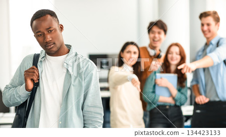 Abuse And Racism. Diverse People Bullying And Whispering Behind Back Of Unhappy Black Man With Backpack Standing Alone In Classroom, Pointing Fingers At Outcast Person, Fake Bad Friends 134423133