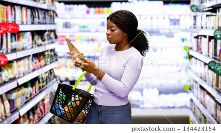 Beautiful African American woman making choice of products at supermarket. Focused female consumer shopping for groceries, selecting food, reading labels at modern mall 134423154