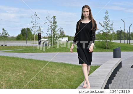 Woman in black dress walking barefoot on pavement in a park on a sunny day. Woman in black dress walking barefoot on pavement in a park on a sunny day. 134423319