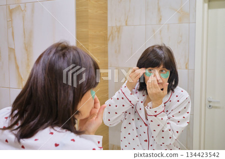 Mature Woman applying green eye patches on her face for skincare routine 134423542