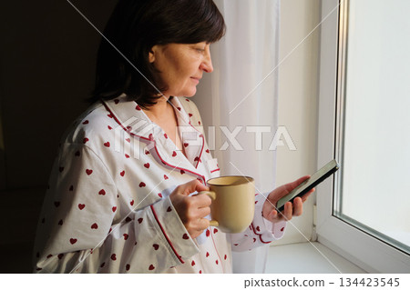 Woman in pajamas drinking coffee and using smartphone near window in morning light 134423545