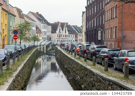 Narrow canal street in Wismar lined with parked cars on both sides beside stone embankment water reflections. Urban oldtown scene shows full parking, historic architecture and everyday city traffic Narrow canal street in Wismar lined with parked cars on both sides beside stone embankment water reflections. Urban oldtown scene shows full parking, historic architecture and everyday city traffic 134423789