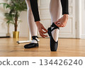 Ballerina puts on pointe shoes on leg in dance class. Classical ballet dancer woman in dance studio. Ballerina putting on tying classical pointe shoes for dance training. Ballet school concept 134426240