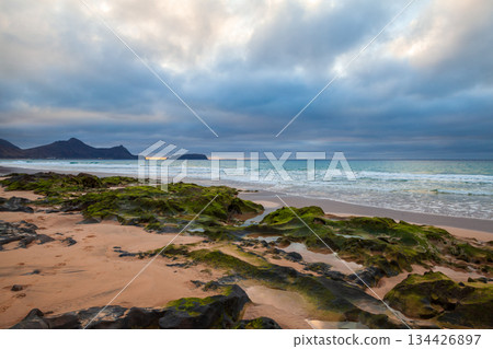 A serene beach scene with dark rocks, soft waves, and a moody, cloud filled sky 134426897