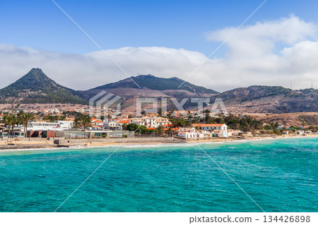 A sunny coastal town sits along a bright turquoise bay, framed by rugged hills A sunny coastal town sits along a bright turquoise bay, framed by rugged hills 134426898
