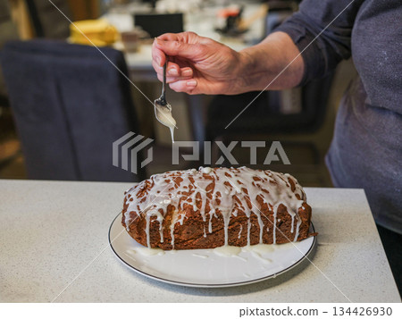 Hand drizzling icing on freshly baked loaf cake on kitchen countertop 134426930