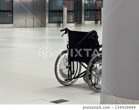 Close view of empty wheelchair positioned beside airport column. Mobility assistance concept, accessibility services, inclusive travel environment, and support infrastructure in public transport. 134426994