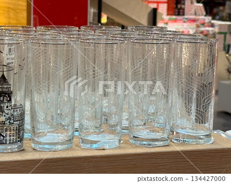 Tall clear drinking glasses arranged on wooden shelf in store. Glassware assortment, retail display, everyday tableware, minimalist design, consumer goods presentation and household utility Tall clear drinking glasses arranged on wooden shelf in store. Glassware assortment, retail display, everyday tableware, minimalist design, consumer goods presentation and household utility 134427000