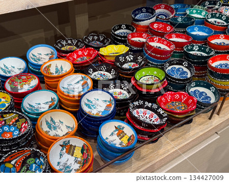 Colorful handmade ceramic Turkish bowls arranged on wooden shelves. Tableware variety, functional art, culinary culture, artisan production, household tradition, decorative ceramics and retail 134427009