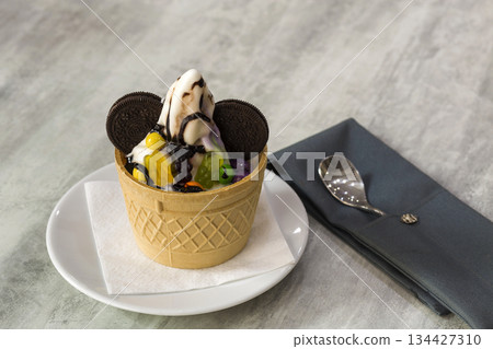 Ice cream dessert in wafer cup with chocolate cookies and creative decoration topping on blurred light copy space background. Ice cream dessert in wafer cup with chocolate cookies and creative decoration topping on blurred light copy space background. 134427310