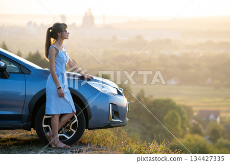 Happy young woman in blue dress standing near her vehicle looking at sunset view of summer nature. Travelling and vacation concept. Happy young woman in blue dress standing near her vehicle looking at sunset view of summer nature. Travelling and vacation concept. 134427335