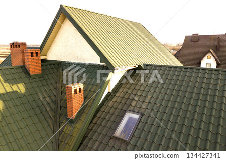 Green metal shingled house roof with attic plastic window and brick chimney. 134427341