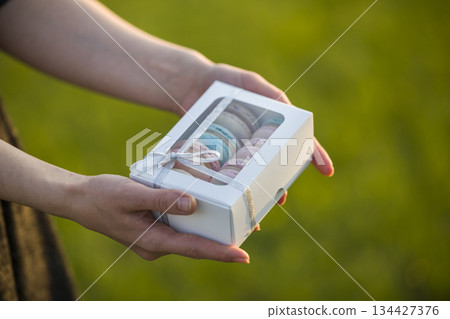 Female hands holding cardboard gift box with colorful pink blue handmade macaron cookies on green blurred copy space background. Female hands holding cardboard gift box with colorful pink blue handmade macaron cookies on green blurred copy space background. 134427376