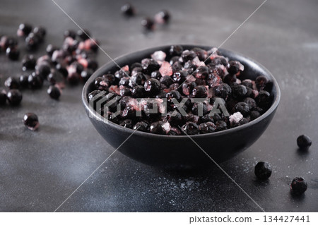 Frozen aronia or chokeberry berries with ice crystals in black bowl on dark stone background, healthy superfood ingredient concept. Raw food Frozen aronia or chokeberry berries with ice crystals in black bowl on dark stone background, healthy superfood ingredient concept. Raw food 134427441