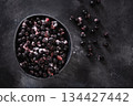 Frozen aronia berries in bowl with scattered berries on dark stone surface, healthy ingredient with copy space. 134427442