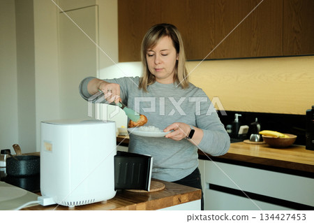 Woman uses tongs to serve food from white air fryer onto plate of rice. Smart air fryer, connected kitchen appliances, modern cooking, healthy eating. 134427453