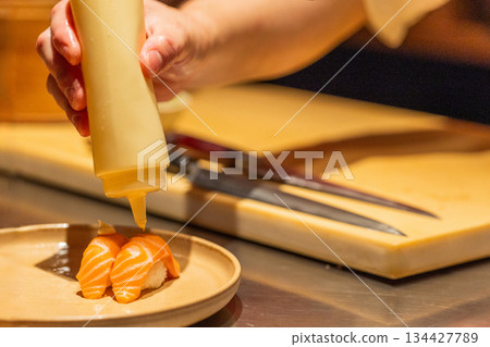 Sushi chef applying sauce to salmon nigiri on wooden countertop 134427789