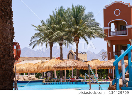 Empty deck chairs under straw shade umbrellas on swimming pool side in tropical resort. Summer vacations and getaway concept Empty deck chairs under straw shade umbrellas on swimming pool side in tropical resort. Summer vacations and getaway concept 134429709