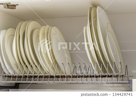 Dish drying metal rack with big nice white and light yellow clean plates on blurred background. Traditional comfortable kitchen. 134429741
