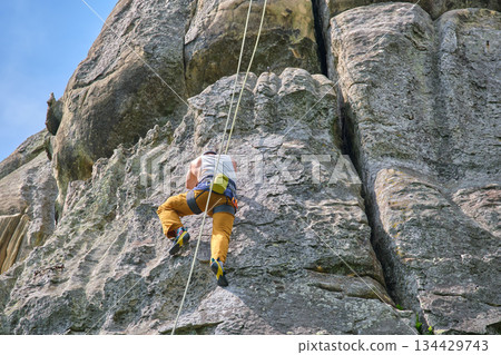 Determined climber clambering up steep wall of rocky mountain. Sportsman overcoming difficult route. Engaging in extreme sports and rock climbing hobby concept. 134429743