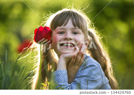 Cute pretty smiling child girl with gray eyes and long hair with bright red tulip flower on blurred sunny green bokeh background. Love to nature concept. 134429760