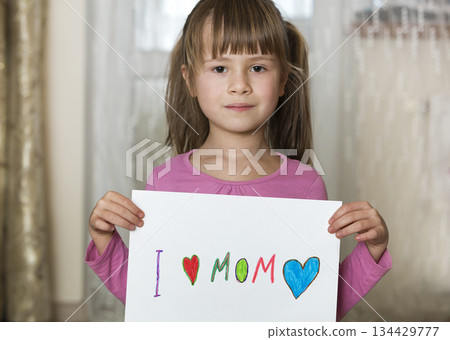 Cute child girl wit sheet of paper with colorful crayons painted words I love Mom. Art education, creativity concept. Cute child girl wit sheet of paper with colorful crayons painted words I love Mom. Art education, creativity concept. 134429777