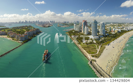 Commercial container ship entering Miami port harbor through main channel near South Beach. Luxurious hotels and residential buildings on waterfront and high skyscraper towers of downtown in distance 134429804