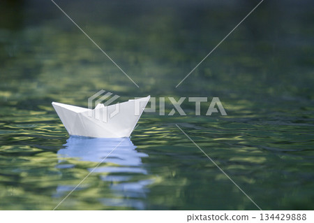 Close-up of simple small white origami paper boat floating quietly in blue clear river or sea water under bright summer sky. Freedom, dreams and fantasies concept, copyspace background. Close-up of simple small white origami paper boat floating quietly in blue clear river or sea water under bright summer sky. Freedom, dreams and fantasies concept, copyspace background. 134429888