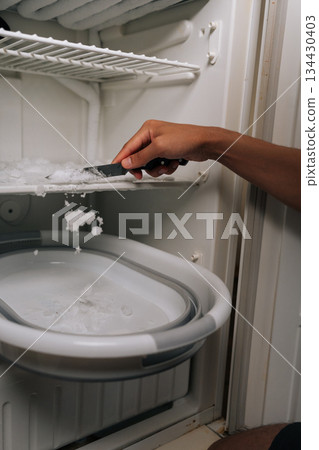 Vertical cropped shot of unrecognizable man defrosting refrigerator, scraping ice buildup with knife and gathering frozen fragments inside basin for cleaning and maintenance. Concept of domestic work. 134430403