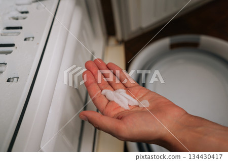 Male hand holding chunk of ice extracted from freezer during defrosting process, with melting ice pooling in containers positioned below, highlighting chore of cleaning and maintenance 134430417