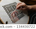 hands carefully stitching a delicate geometric lace pattern on white linen stretched in an embroidery hoop 134430722