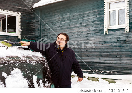 Snow brushing car windshield using automotive brush, person clears vehicle surface beside wooden house wearing winter clothing. 134431741