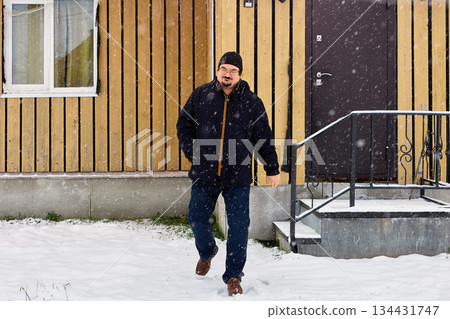 Emerging from wooden building with vertical siding, warmly dressed man over fifty moves into snowy yard amid falling snow during winter daylight. Emerging from wooden building with vertical siding, warmly dressed man over fifty moves into snowy yard amid falling snow during winter daylight. 134431747