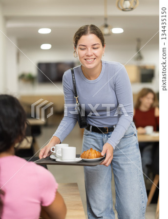 Smiling young girl putting coffee and croissant on table in self-service cafeteria Smiling young girl putting coffee and croissant on table in self-service cafeteria 134435150