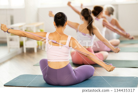 Women of different ages sit on mats and do pilates exercises with an expander in an gym Women of different ages sit on mats and do pilates exercises with an expander in an gym 134435317
