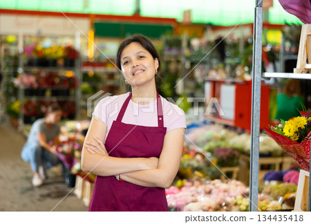 Portrait of smiling young woman gardener standing at greenhouse with flowers on background 134435418