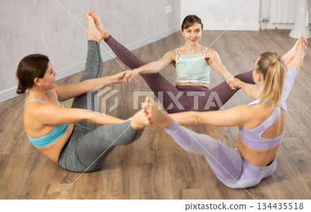 Positive young sports women making fun and sitting in front of each other, touching feet and holding hands together in yoga studio Positive young sports women making fun and sitting in front of each other, touching feet and holding hands together in yoga studio 134435518