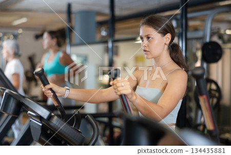 Slender athletic girl during cardio exercises on elliptical simulator in fitness center. 134435881