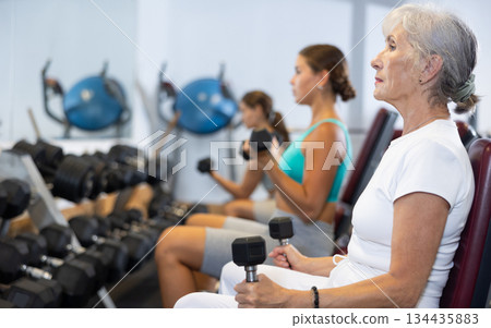 Slender senior woman sit and trains muscles of arms and back using dumb-bell shaped figure weighting 134435883