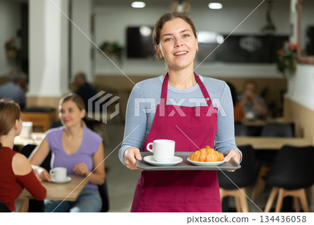 Female cafe employee carries coffee and croissants on tray Female cafe employee carries coffee and croissants on tray 134436058