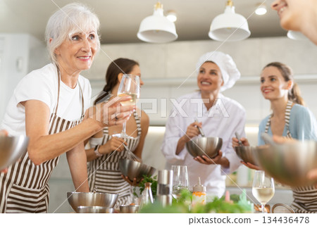 Aged woman holding wine and talking to other cooking course members whipping souce in bowls 134436478