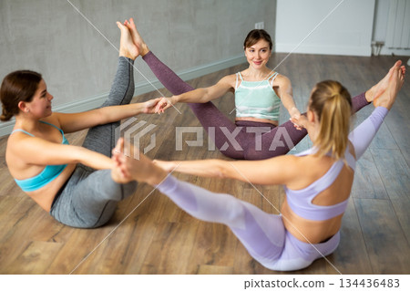 Three smiling women practicing upright seated angle pose of yoga in light fitness room Three smiling women practicing upright seated angle pose of yoga in light fitness room 134436483