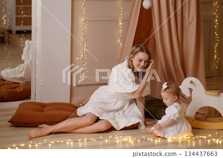 Mother playing with baby girl on floor in cozy kids room with wooden horse and soft pillows 134437663