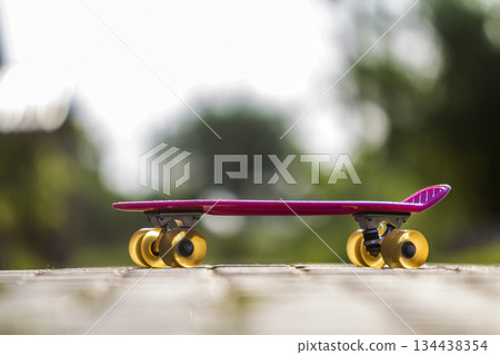 Close-up of child plastic pink skateboard isolated on pavement against bright white and green blurred bokeh background. Sport, recreation, fun and plays concept. 134438354