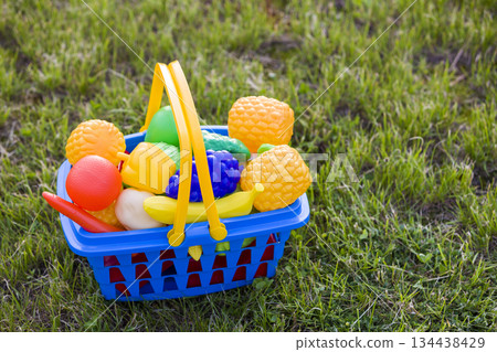 Bright plastic colorful basket with toy fruits and vegetables outdoors on sunny summer day. 134438429