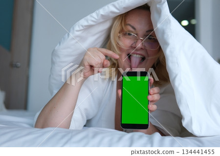 Middle aged woman under blanket sticking tongue out smiling holding smartphone with green screen showing potential advertising or digital content. 134441455