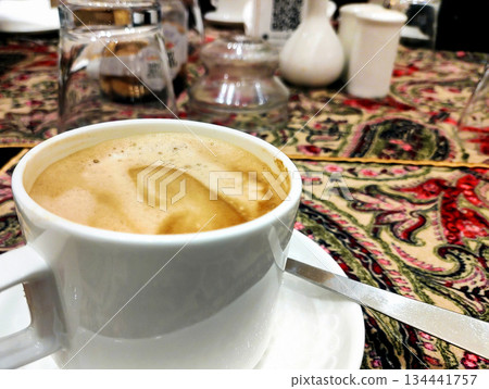 Close-Up of Frothy Coffee in White Cup on Traditional Tablecloth Close-Up of Frothy Coffee in White Cup on Traditional Tablecloth 134441757