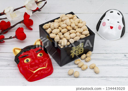 Setsubun soybeans in a box, red demon and Okame masks, and red and white plum branches placed on a white wood background 134443020