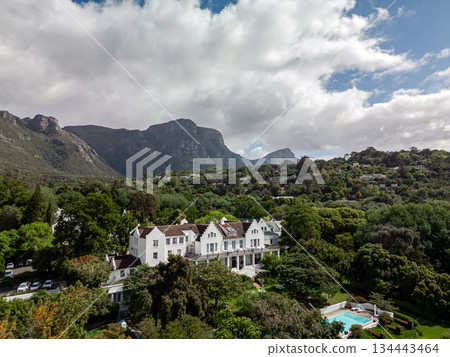 Large white estate with a pool nestled in a lush green valley, surrounded by majestic mountains. Scenic landscape with elegant architecture for travel and luxury brochures. 134443464