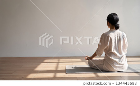 陽光明媚地灑進一間乾淨的房間，一位女士背對著鏡頭，在瑜珈墊上冥想。 134443688